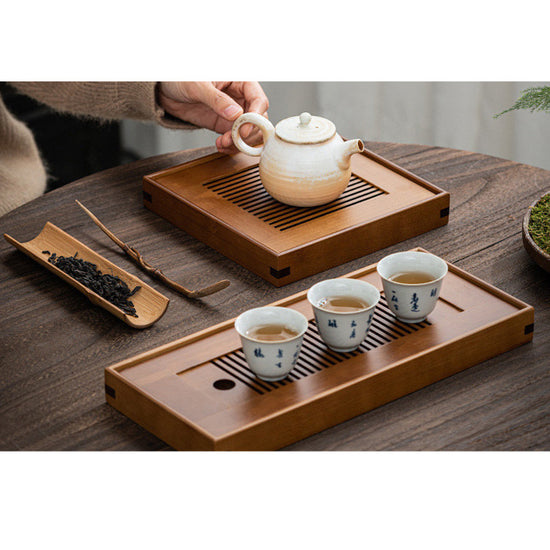 Two bamboo tea trays next to each other placed on wooden table with tea tools and porcelain teaware